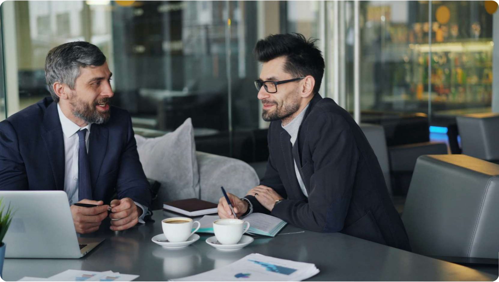 Two advisers in discussion across a table in a modern office, reviewing documents over coffee.
