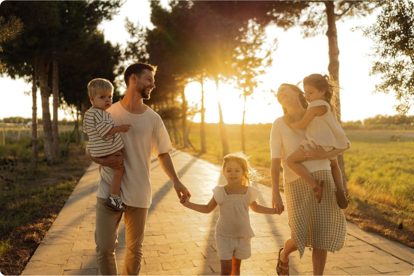 A family of four walking hand in hand along a tree-lined path at sunset.
